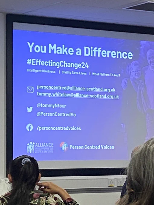 A projection screen at the HCSW / CSW Conference displaying a presentation slide titled "You Make a Difference" with the hashtag "#EffectingChange24". The slide includes themes of "Intelligent Kindness | Civility Save Lives | What Matters To You?" and contact information: personcentred@alliance-scotland.org.uk, tommy.whitelaw@alliance-scotland.org.uk, Twitter handles @tommyNtour and @PersonCentredVo and a Facebook page /personcentredvoices. Logos for Alliance Health and Social Care Alliance Scotland and Person Centred Voices are at the bottom. In the foreground, two audience members with their backs to the camera are visible, one with long dark hair in a ponytail and another with grey hair.