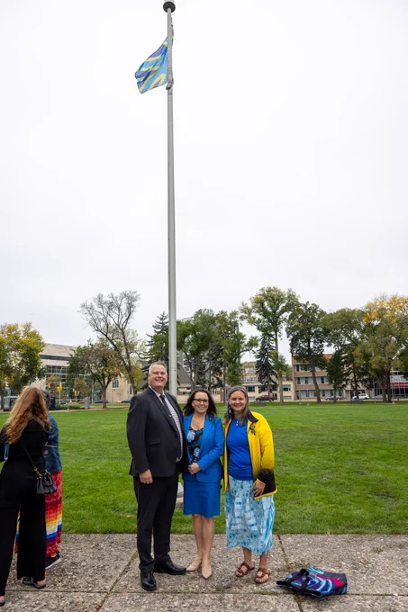 MLAs Wayne Balcaen and Jodie Byram with the Manitoba Deaf Association's Shawna Joynt in front of the Deaf flag raised at Memorial Park.