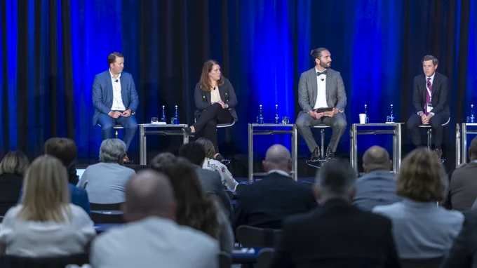 Four panelists seated on stage in front of an audience at a conference, consisting of two individuals on the left casually dressed and two on the right in business attire.