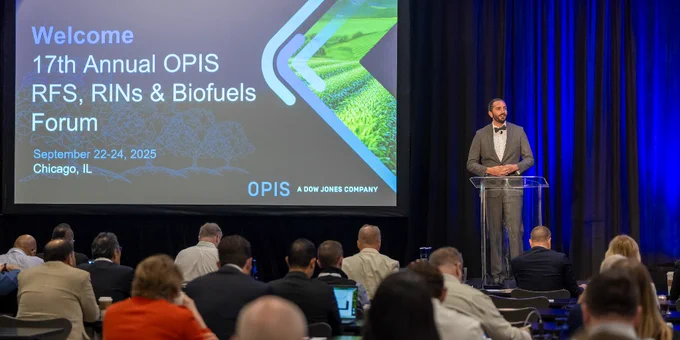 A person presenting at the 17th Annual OPIS RFS, RINs & Biofuels Forum in Chicago, visible on the stage with a large projection screen behind displaying the event details. Audience members are seated, facing the presenter.