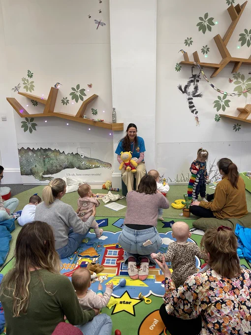 A woman in a blue t-shirt is leading a Bookbug session for parents and small children in a library.