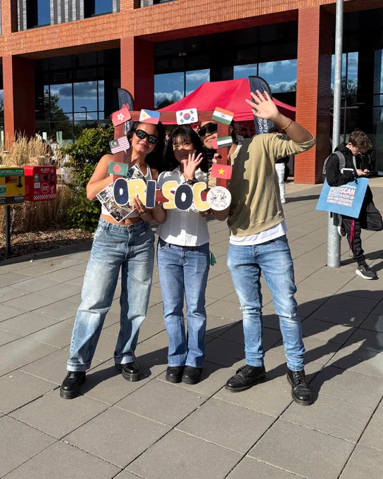 Three individuals stand outside a building promoting the 'Ori-Soc' with handmade signs and flags.