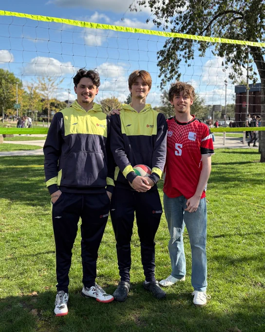Three people standing on a grassy field with a volleyball net above them, dressed in athletic gear.