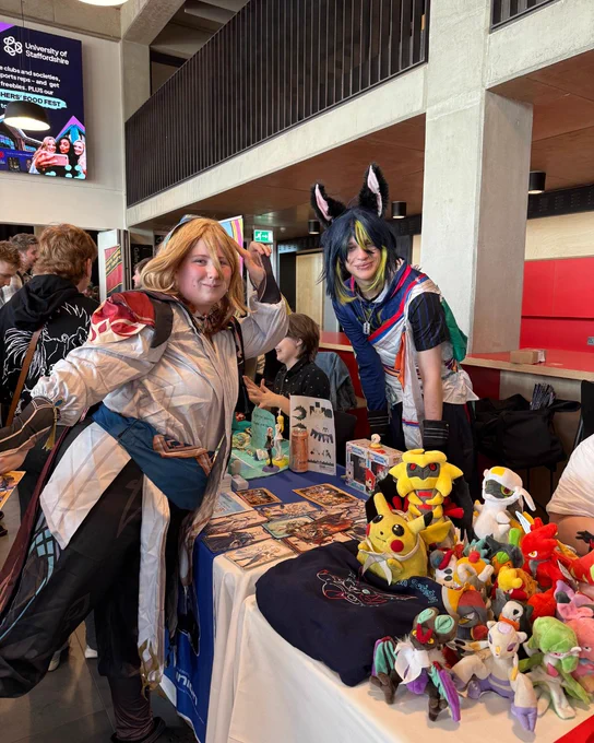 Two individuals at a convention booth with one dressed in a fantasy costume and the other wearing a hoodie with animal ears. The table in front of them is covered with various plush toys, including multiple Pokémon characters.