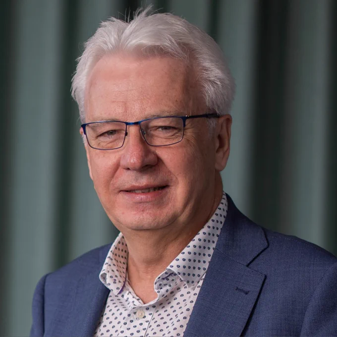 Jim McLaughlin wearing glasses, a blue blazer and a white shirt with blue polka dots against a green curtain background.