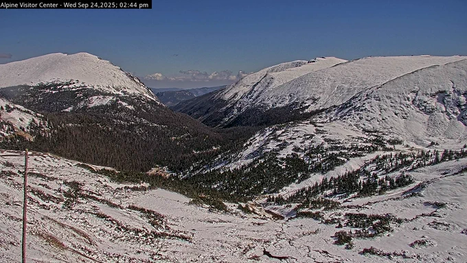 An afternoon view from the Alpine Visitor Center webcam. Snow covers the landscape below Alpine Visitor Center. The sky is blue and clear.