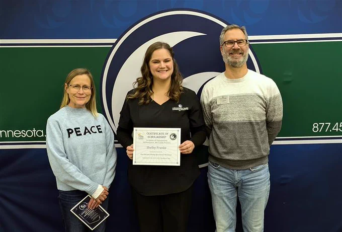 Three individuals standing in front of a banner with the Minnesota State Community and Technical College's logo, smiling as the person in the center holds a Certificate of Commendation.
