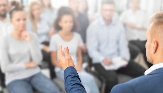 Business professional giving a presentation or training session to a seated audience, with focus on the speaker’s hand gesture and blurred attendees in the background.