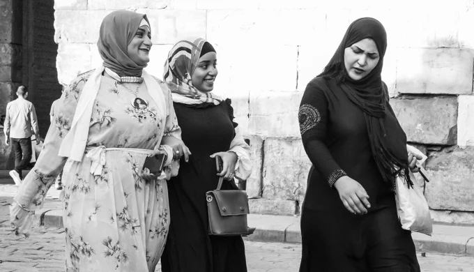 Black and white photo of three women in hijabs walking together on a cobblestone street, two smiling and chatting while the third looks down thoughtfully.