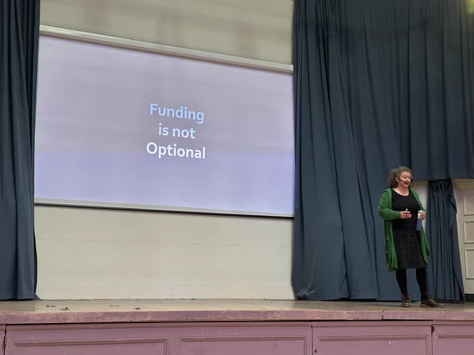 Juliette in front of a slide that says “Funding is not optional”.