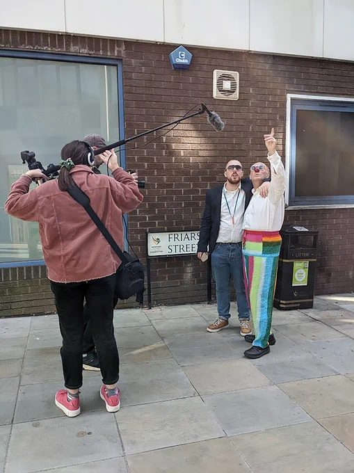 Two people standing close together raising an arm and looking up, while a crew films them with a boom mic and camera.