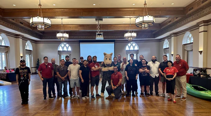 Group of people posing for a photo in a large hall with a mascot in the center, at the University of Oklahoma.