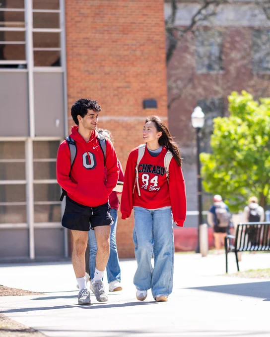 Two students wearing red sweatshirts walk and talk on a sunny campus pathway, with other students visible in the background.