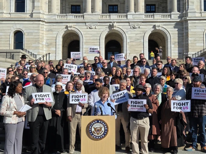 Photo depicts Senator Tina Smith at the podium, surrounded by multiracial, multifaith faith leaders.