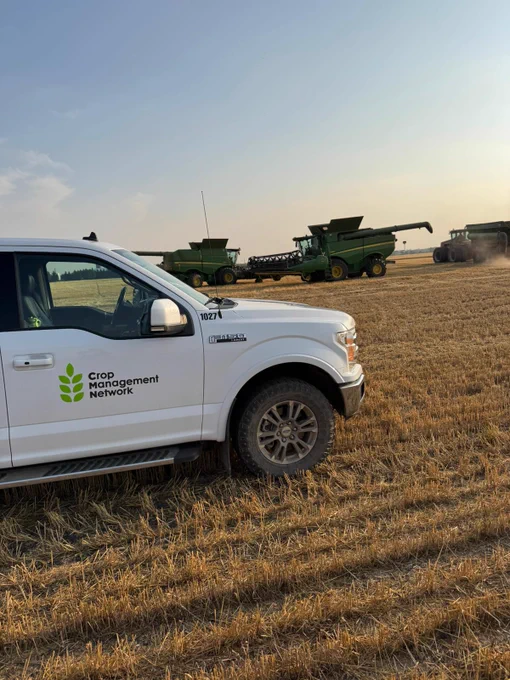 A white pickup truck with "Crop Management Network" logo parked in a field, with two green John Deere combines in the background under a hazy sky.