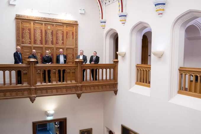 Six men in formal attire stand on a wooden balcony inside a historic building with ornate carved panels behind them.