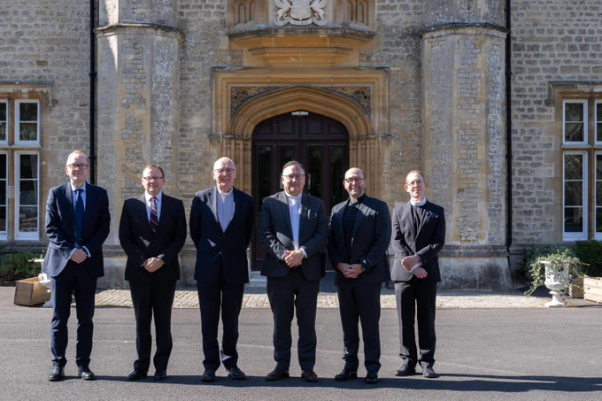 Six men dressed in formal suits stand in a row outside a historic stone building with an ornate arched doorway.