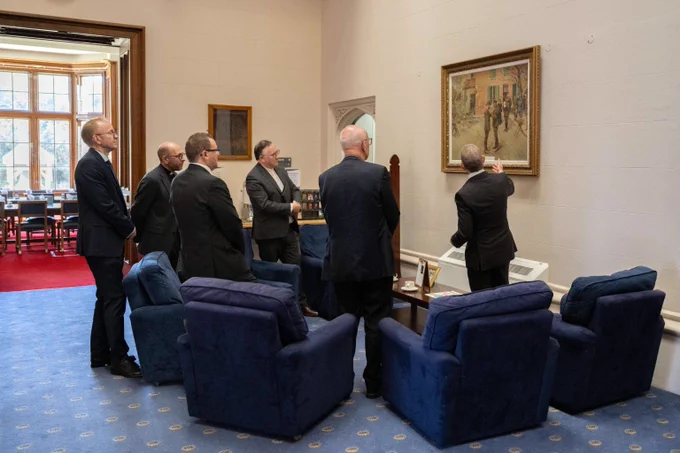 A group of six men in suits attentively observe a framed painting in a formal room with blue armchairs and carpet.
