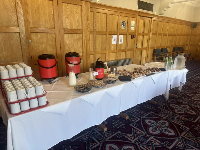 A picture of a long table with white table cloths. There are two large red flasks with tea and coffee, white cups, milk bottles and pastries on the table.