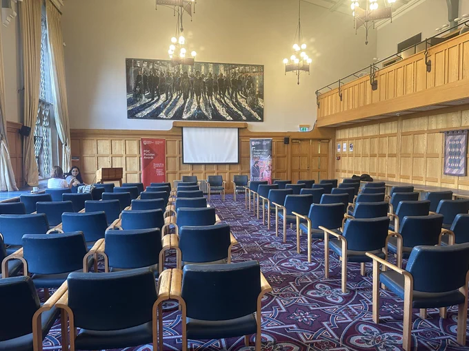 Picture of the Council Chamber, Lanyon Building, QUB with chairs in rows/theatre style in the room. A painting of women coming out of shadows is situated above a white screen at the front of the room. A PDC Postdoc Awards pop up stand in red and a PDC pop up stand with an image of a female and male walking beside each other smiling are located at each side of the screen with 3 chairs in the middle for panellists.