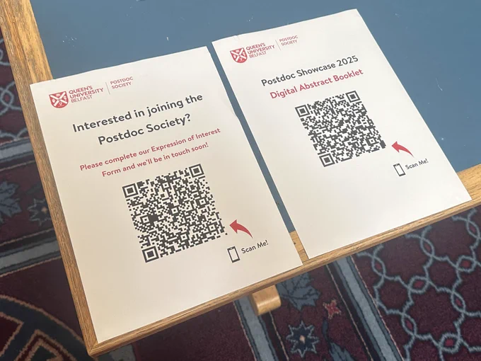 Two signs on white A4 paper with QR codes that take you to a register of interest form to join the Postdoc Society and the Postdoc Showcase 2025 digital abstract booklet.
