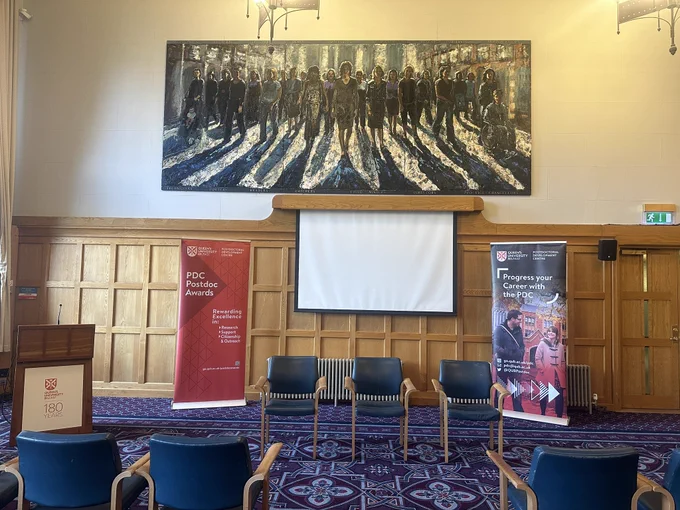 Picture of the front of the Council Chamber, Lanyon Building, QUB with a painting of women coming out of shadows above a white screen. A PDC Postdoc Awards pop up stand in red and a PDC pop up stand with an image of a female and male walking beside each other smiling are located at each side of the screen with 3 chairs in the middle for panellists.