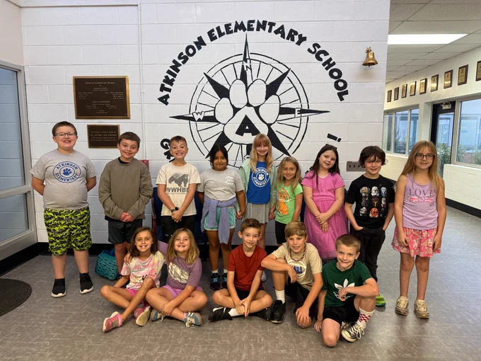 14 students gather together in the lobby. 5 students sit on the ground and the rest stand. they are smiling at the camera. the atkinson compass mural is in the background painted on the wall. 