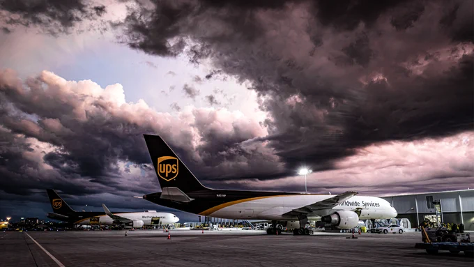 Two UPS planes on the tarmac under a sky dominated by thick, dark clouds tinged with hues of pink and purple.