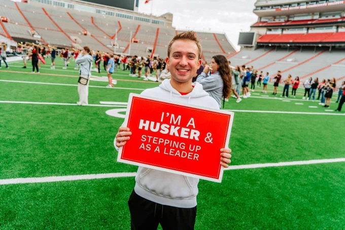 Tanner with a red sign that reads, "I'm Husker and stepping up as a leader," while standing in Memorial Stadium as the Cornhusker Marching Band practices.