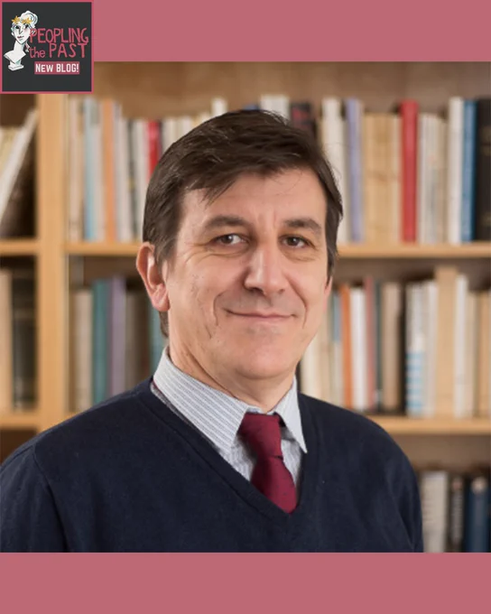 A headshot photo of Goran. He is wearing a shirt and tie, with a baby blue sweater/ He stands in front of a bookcase filled with books and is smiling.