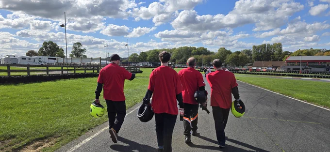Four people in red shirts walking down a racetrack, each carrying a helmet.