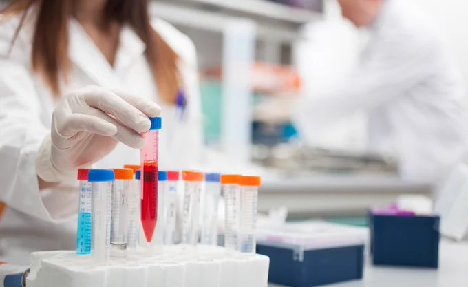 A person in a lab coat and gloves holds a test tube filled with red liquid. The test tube is in a foam holder alongside several other capped vials, each with colored tops. The background is a blurred laboratory setting, with another person in a white coat working.