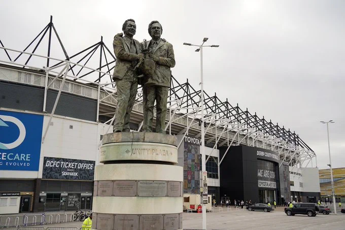 Statue of Brian Clough and Peter Taylor shaking hands on a round pedestal outside Pride Park Stadium, with the stadium's West Stand and cloudy skies in the background.
