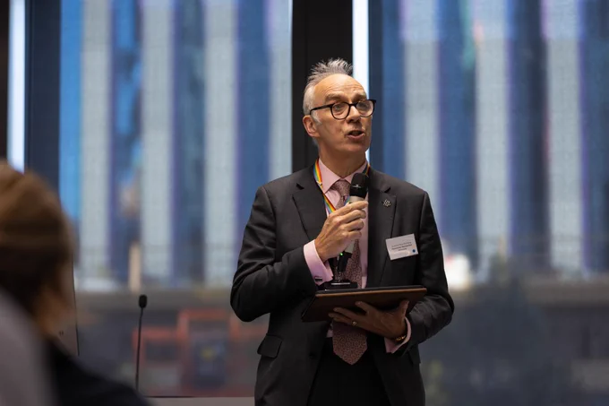 A person holding a tablet and speaking at a conference podium, with a cityscape visible through the large window in the background.
