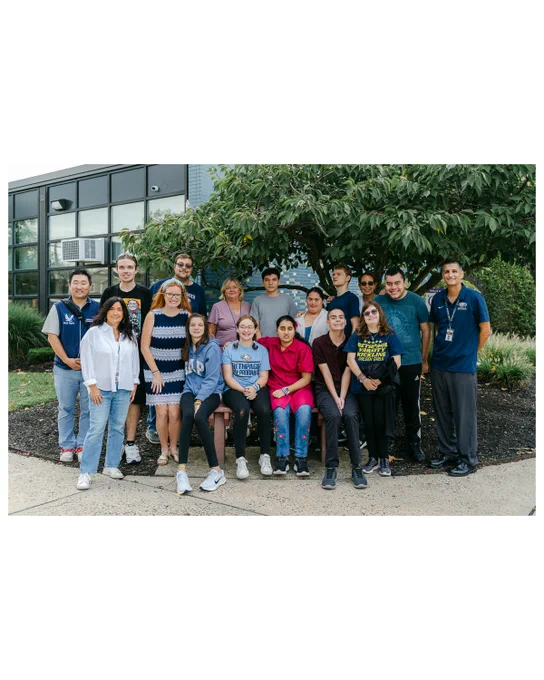 A group of Bethpage High School ACE students and staff pose together outside the school building. The students are seated on a bench and standing around it, smiling at the camera, with a tree and school windows in the background.