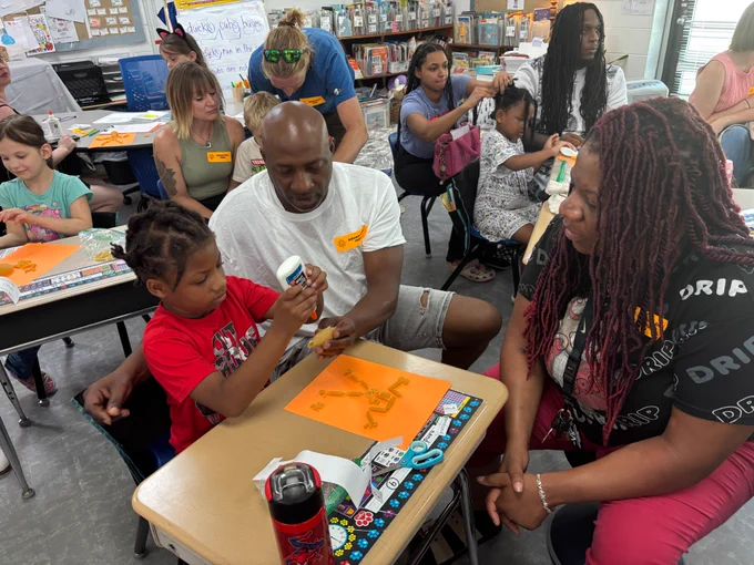 first grade boy sits at his desk next to his dad and across from his mom. they are using glue to create skeletons out of uncooked noodles