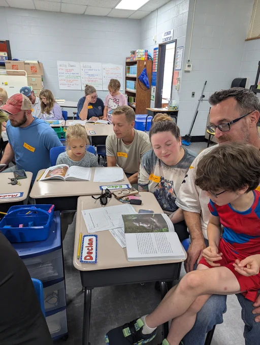 group of students and parents sit at a u shape of desks in mrs. jackson's third grade class. they are all actively looking at an open book in front of them and working together