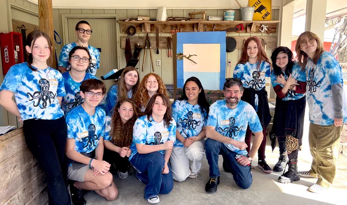 A group of 15 people poses together in a workshop setting, wearing matching blue tie-dye shirts featuring an octopus design. Behind them is a painted backdrop depicting a simple beach scene. The group appears to be engaged and cheerful, conveying a sense of camaraderie.