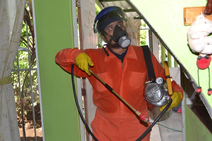 A person wearing protective gear, including an orange suit, yellow gloves, a face mask, and a face shield, sprays insecticide on an indoor wall using a metal canister sprayer as part of an indoor residual spraying activity.