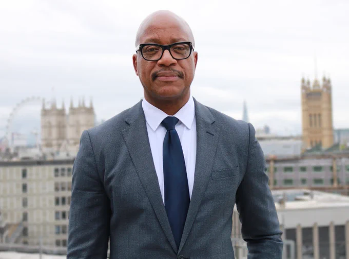 Professor Kevin Fenton wearing a black suit, white shirt and black tie. He is looking towards the camera and smiling.