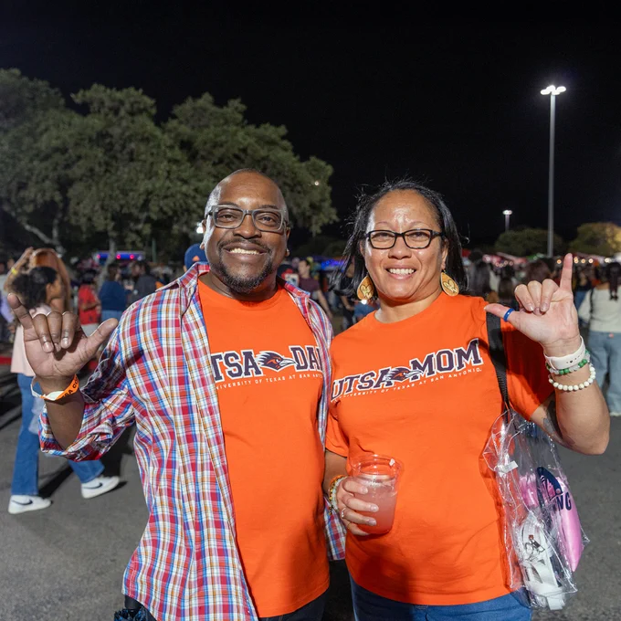 Two individuals wearing "UTSA" attire are happily posing at a night event, each making a Birds Up hand sign.
