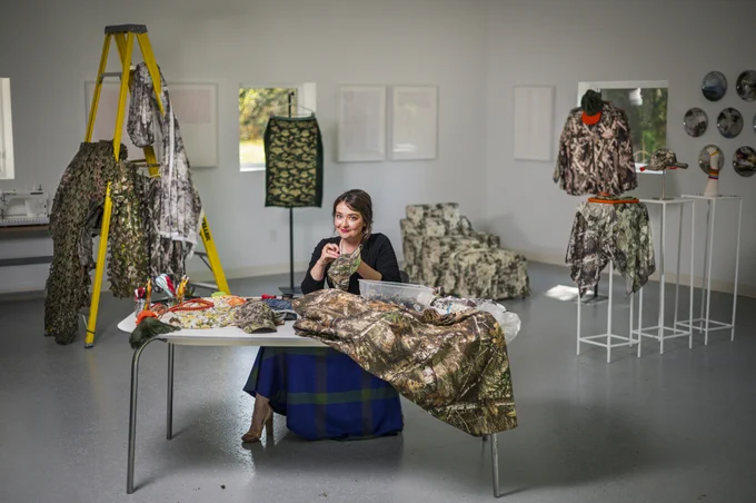 Sophia Brueckner sitting at a table covered with camouflage patterned materials and garments in her home studio decorated with similar themed artworks. A ladder and additional camouflage items are displayed around the room.