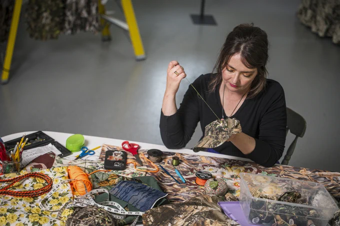 Sophia Brueckner sits at a worktable covered with colorful patterned fabric, threads and tools, diligently embroidering a camo print baseball cap.