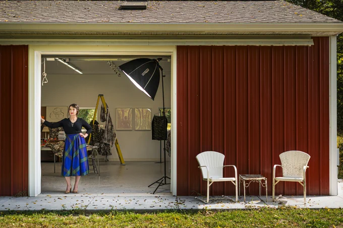 Sophia Brueckner standing in her open garage art studio with artwork, materials and furniture around. Two white chairs are placed outside the garage.
