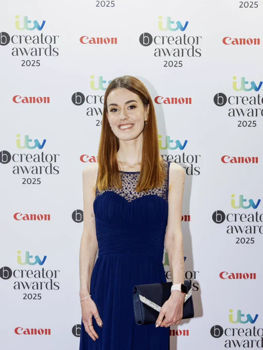 Me smiling on a red carpet infront of a board with the itv bcreator awards 2025 logo on it. I am wearing a long blue dress and holding a blue clutch bag.