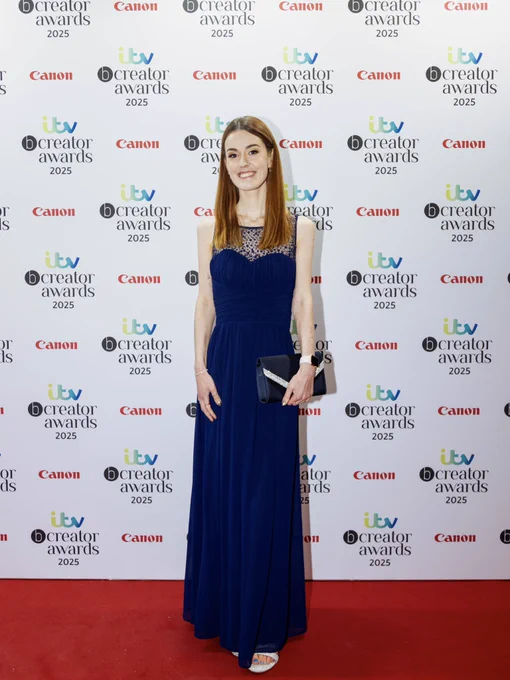 Me smiling on a red carpet infront of a board with the itv bcreator awards 2025 logo on it. I am wearing a long blue dress and holding a blue clutch bag.
