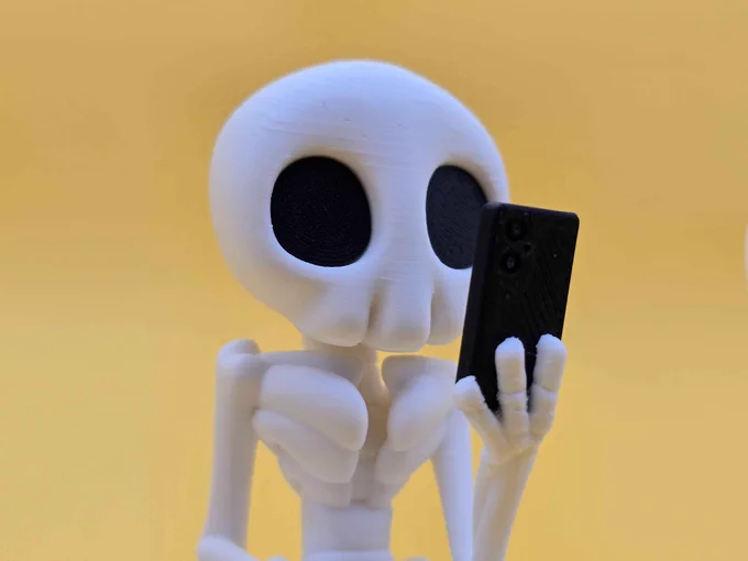 Close-up of a 3D printed Skeleton Hazy figure staring at a black smartphone. Its smooth white bones and large black eye sockets stand out sharply against a warm yellow background.
