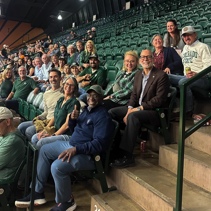 A group of CSU fans sits together in the stands at Moby Arena during a volleyball game. The crowd smiles and looks toward the camera, showing team spirit surrounded by green seats and arena lighting above.