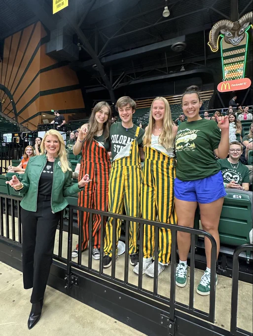 President Amy Parsons stands courtside with four students in the stands at a Colorado State University volleyball game. The group smiles and makes the CSU “ram horns” hand sign in Moby Arena, with other fans and the Ram mascot sign visible in the background.