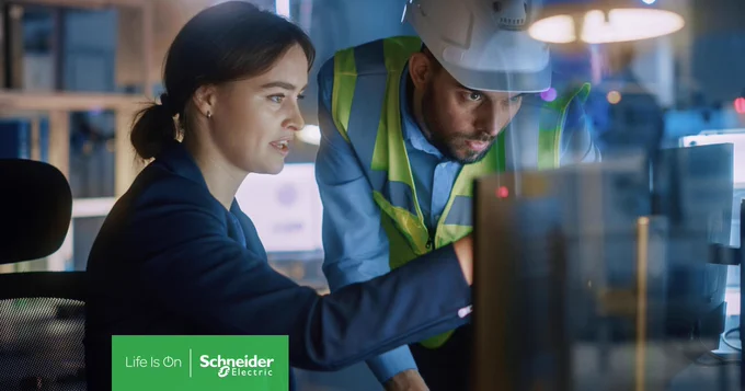 Eine Frau sitzt vor einem Computer. Daneben steht ein Mann mit Schutzhelm und Warnweste. Beide schauen auf den Computerbildschirm. 